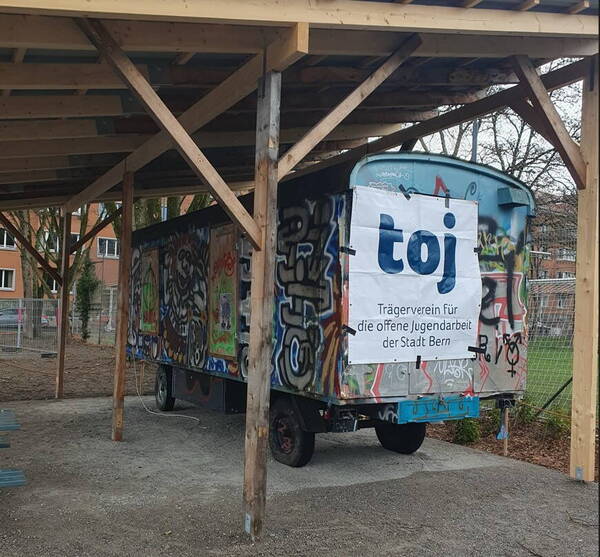 Unter einem Holzunterstand steht der Bauwagen. Er ist voller Graffiti und an der einen Seite ist eine Plache mit dem toj Logo fixiert. Unter einem Holzunterstand steht der Bauwagen. Er ist voller Graffiti und an der einen Seite ist eine Plache mit dem toj Logo fixiert.
