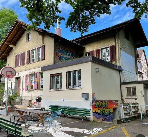 Das Jugendhaus Hopfenrain von aussen. Es hat Bänke, Tische, ein Griff, ein Basketballkorb  und ein Graffiti einer Person am Boden. An einer Wand hat es ein Graffiti auf dem Modi*treff steht.